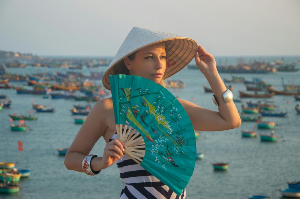 Woman wearing traditional hat holding a vibrant fan at bustling harbor with colorful boats.