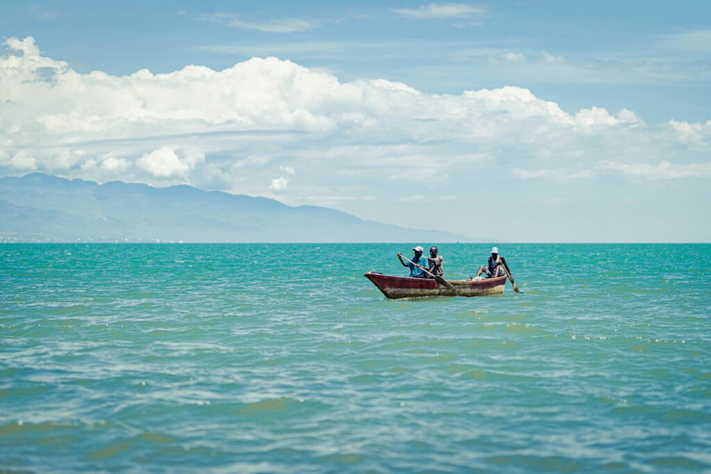Three individuals in a wooden boat paddling through clear turquoise ocean under a bright sky.