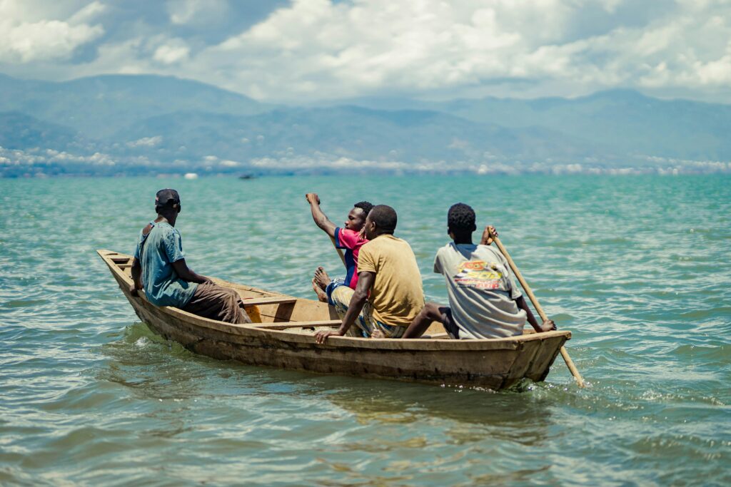 Four men rowing a wooden boat on a serene lake with mountains in the background on a sunny day.