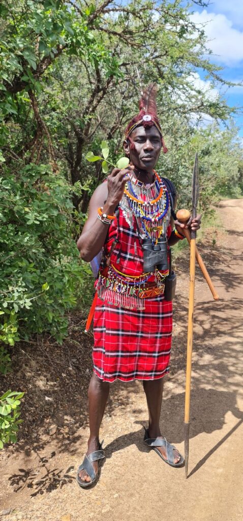 A Maasai warrior in traditional attire holding a spear and fruit in a natural setting.