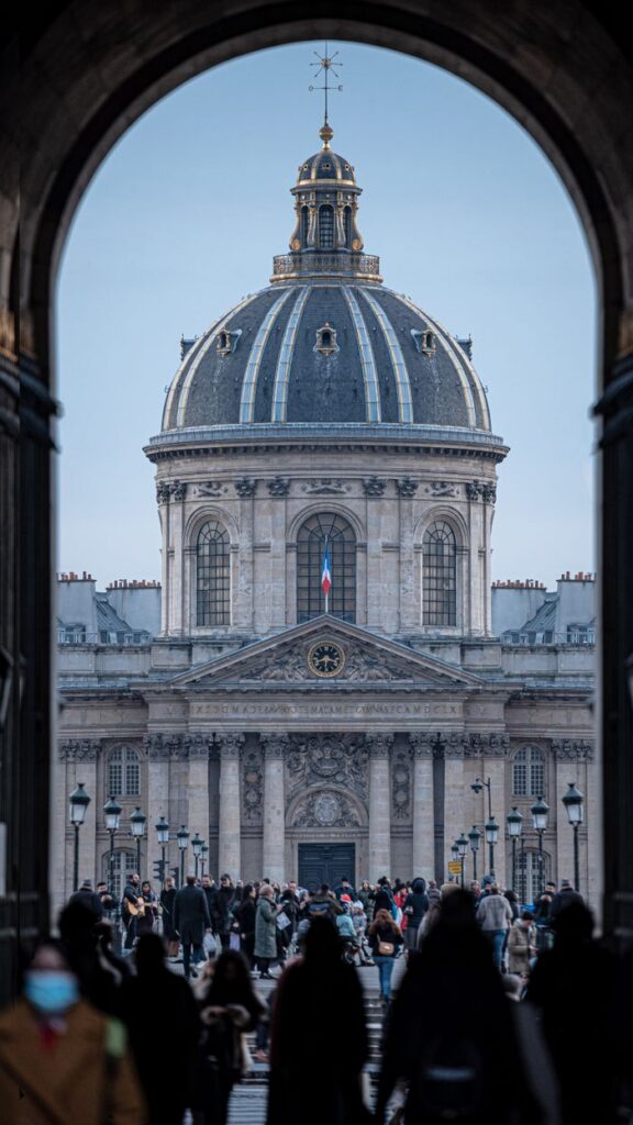 Iconic view of the Institut de France in Paris framed by an arch, capturing the historic facade and a bustling crowd.