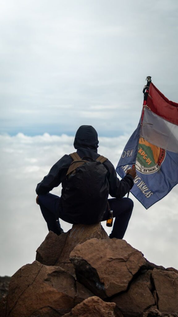 A man holding a flag sits on a rocky mountain peak, overlooking the clouds in Indonesia.