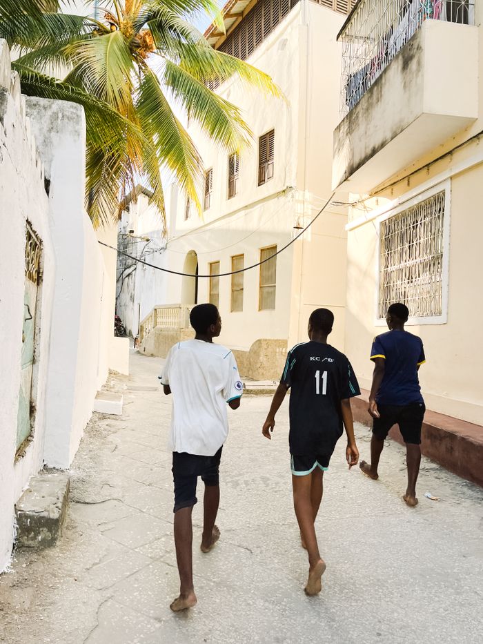 Three teenagers walk down a sunlit urban street in Tanzania with palm trees and buildings surrounding them.