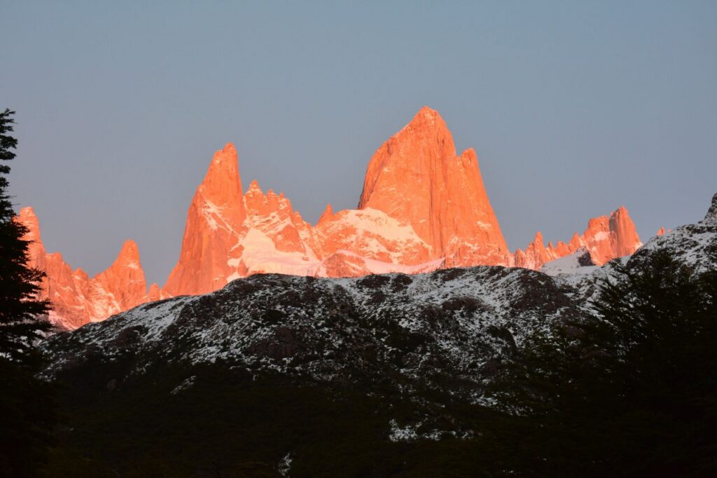 Sunset casts a warm glow on snowcapped jagged mountains, creating a dramatic scene.