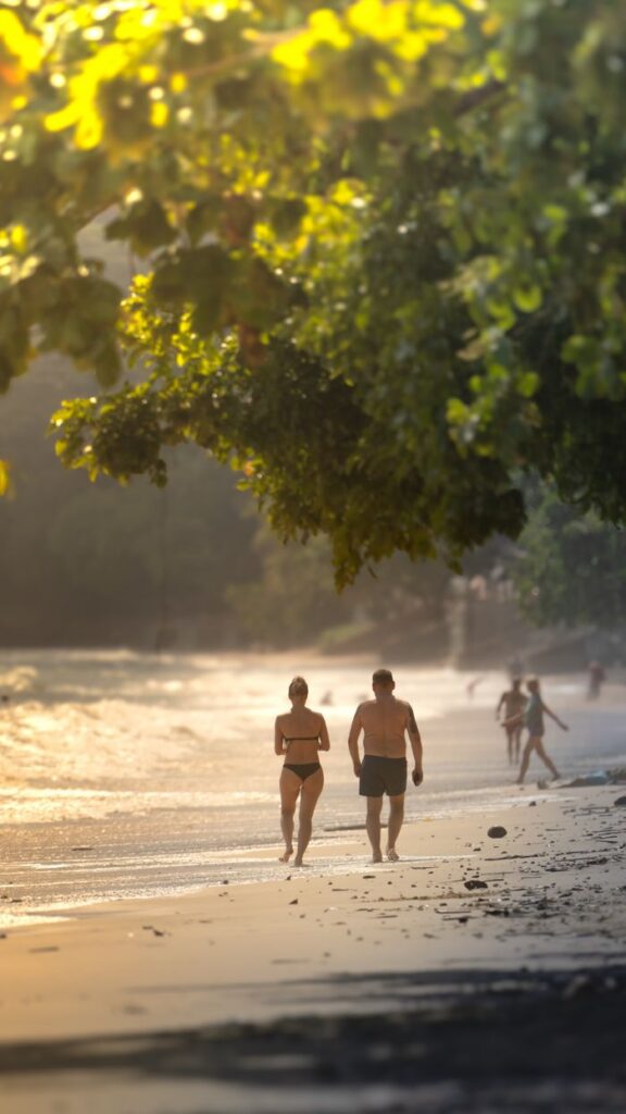 A couple walks along a serene Thai beach at sunset, surrounded by lush greenery and golden sands.