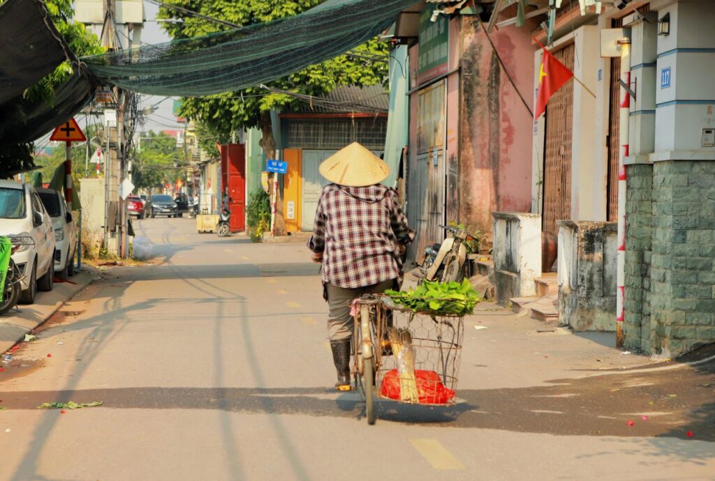 A local cyclist with a traditional conical hat rides through a vibrant Vietnamese village street.